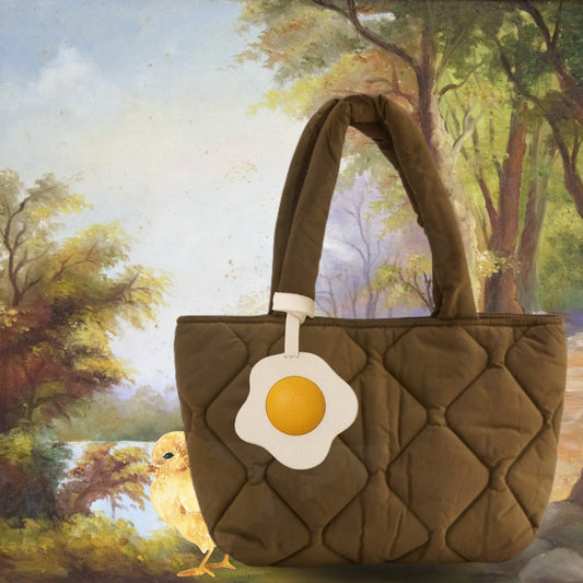 Breloque œuf en cuir fixée à un sac matelassé brun, mise en scène poétique avec un poussin et un décor champêtre.