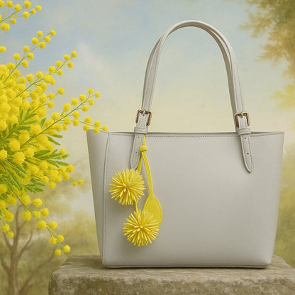 Bijou de sac Pomponnette en cuir jaune, inspiré du mimosa, suspendu à un sac gris sur fond de pierre, avec un bouquet de fleurs de mimosa — accessoire LaMartine.